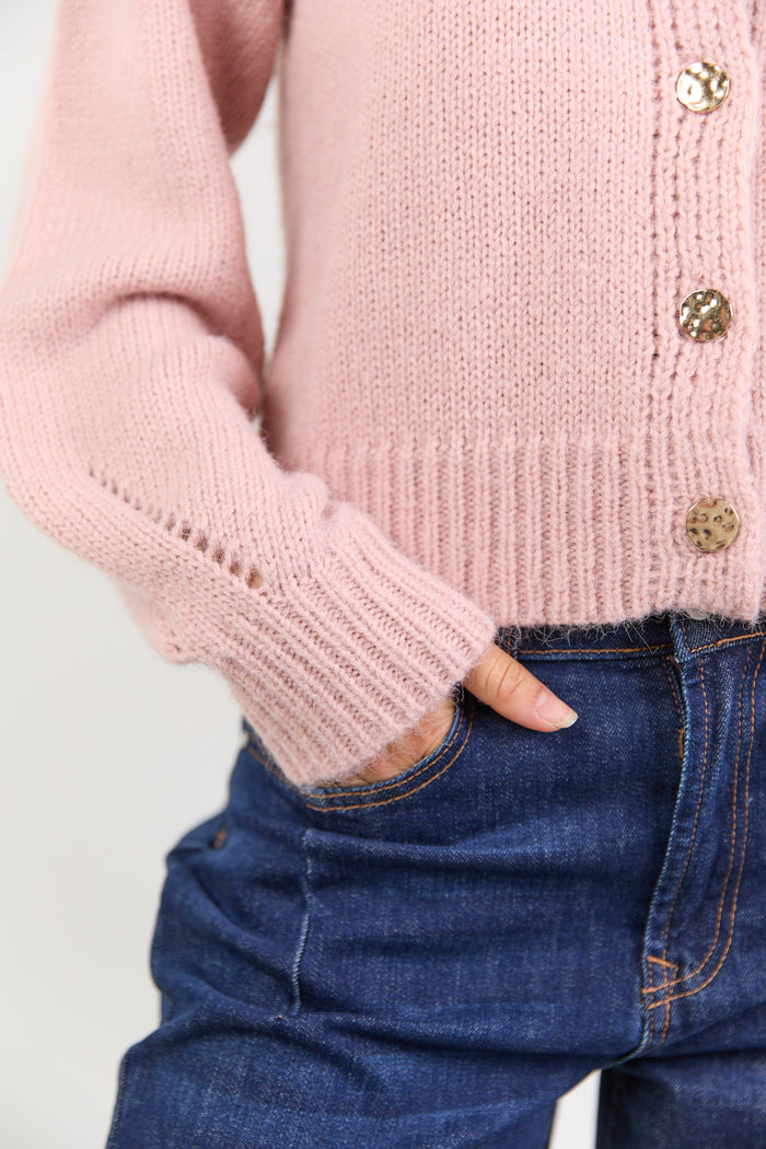 Pink knitted cardigan with gold buttons worn by a person, with a focus on the sleeve and hand.