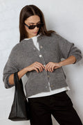 Woman wearing a gray cardigan over a white shirt, holding a black bag against a light background