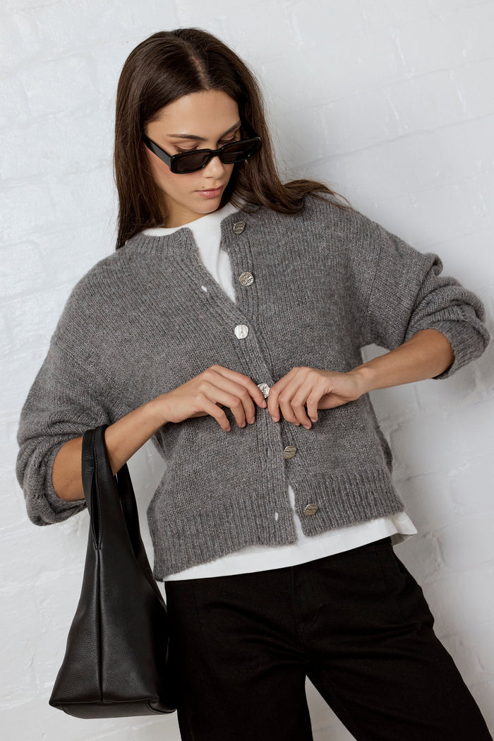 Woman wearing a gray cardigan over a white shirt, holding a black bag against a light background