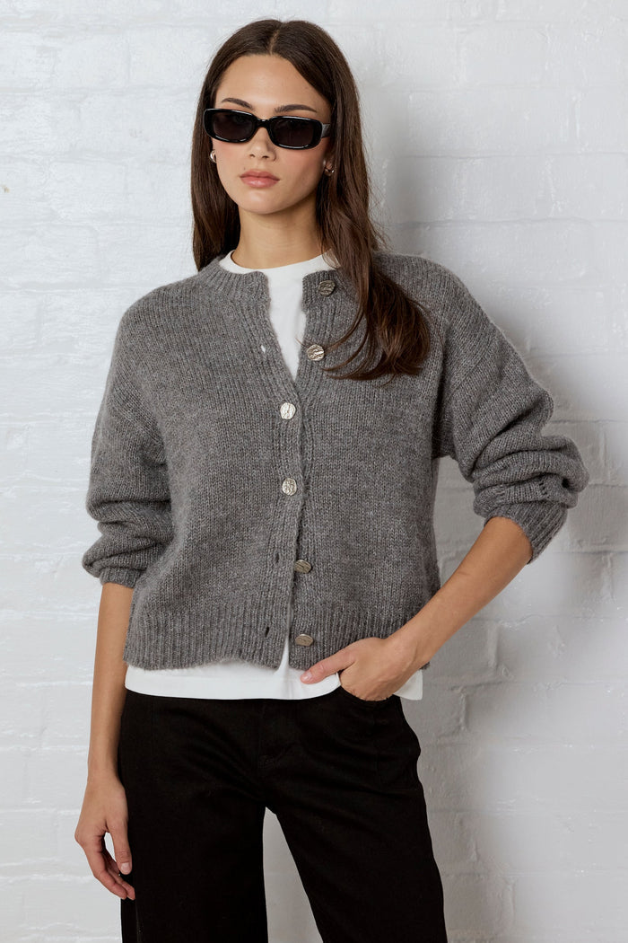Woman wearing a gray cardigan, white shirt, and black pants against a white brick wall.