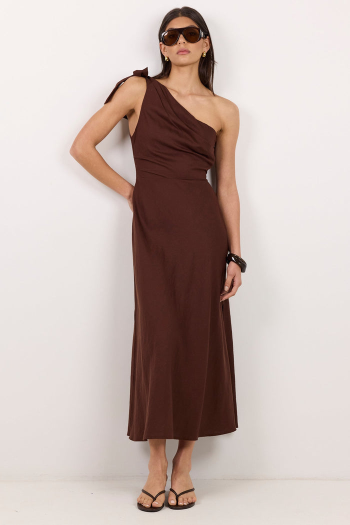 A woman modeling a dark brown one-shoulder midi dress with a draped neckline, standing against a plain white studio background.