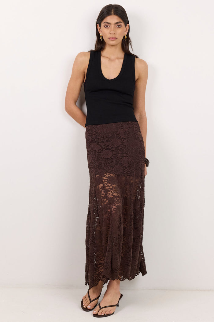 A woman stands against a plain white studio wall wearing a black sleeveless top and a long dark brown lace maxi skirt with a draped side detail.