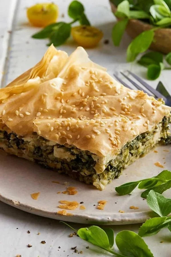 A square slice of layered phyllo pastry pie with a green spinach and feta filling, topped with sesame seeds, served on a light ceramic plate with a fork and knife.