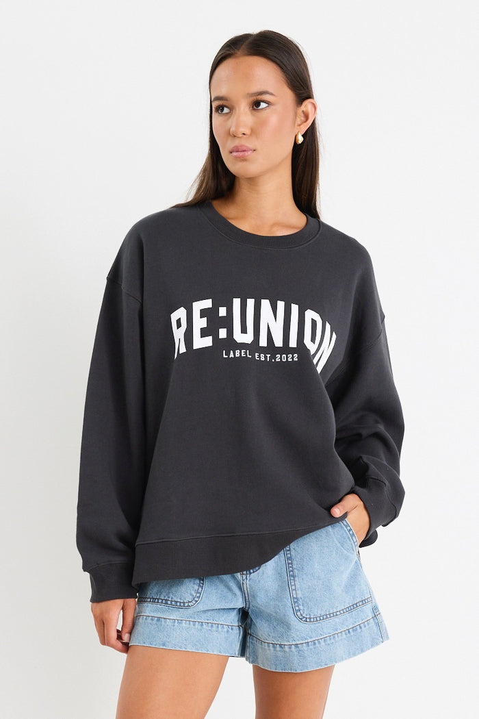 Woman wearing a charcoal grey oversized sweatshirt with "RE:UNION" printed across the chest, styled with light blue denim shorts.