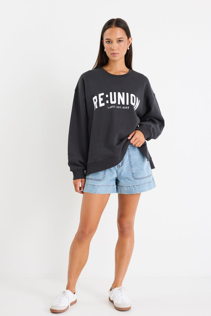 Woman wearing a charcoal grey oversized sweatshirt with "RE:UNION" printed across the chest, styled with light blue denim shorts.