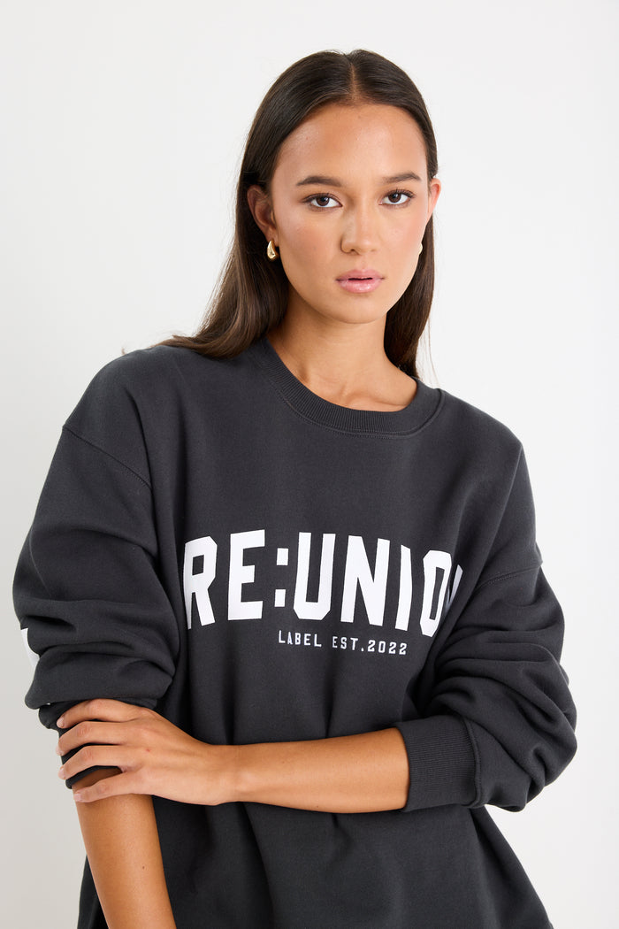 Woman wearing a charcoal grey oversized sweatshirt with "RE:UNION" printed across the chest, styled with light blue denim shorts.