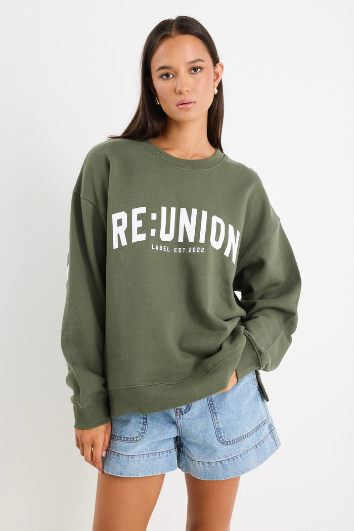 Woman wearing a khaki green oversized sweatshirt with "RE:UNION" printed across the chest, styled with light blue denim shorts.