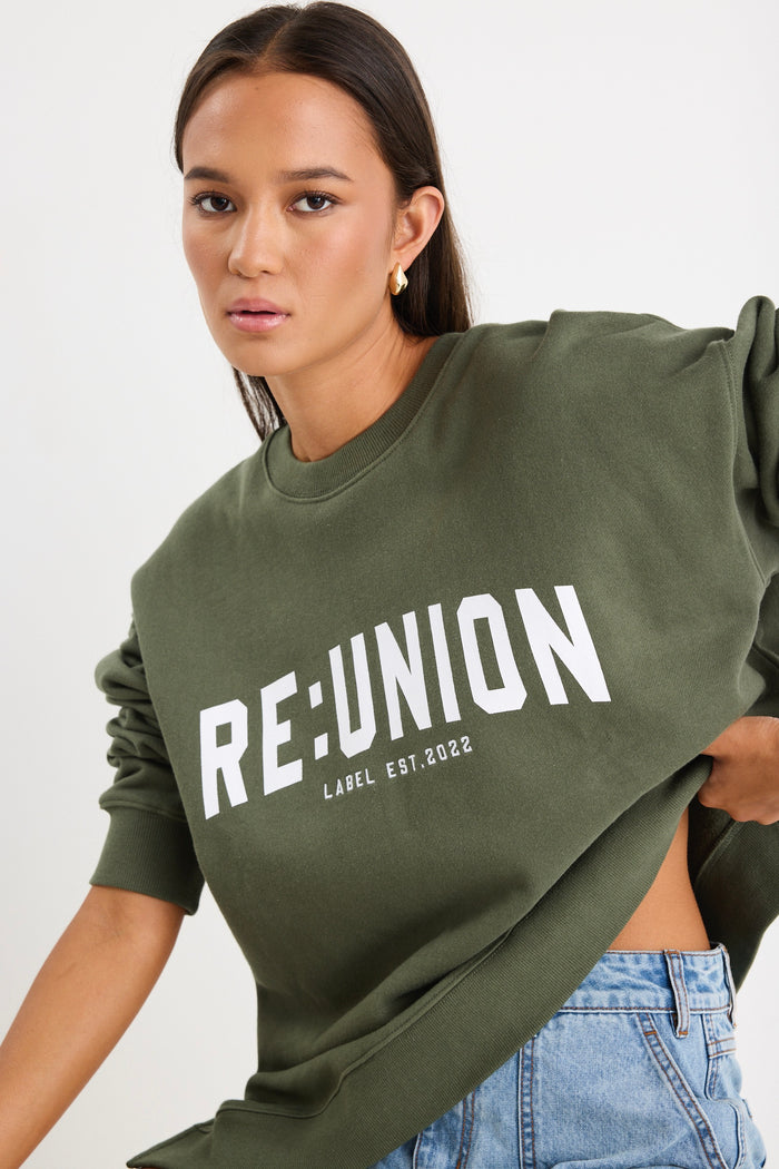 Woman wearing a khaki green oversized sweatshirt with "RE:UNION" printed across the chest, styled with light blue denim shorts.