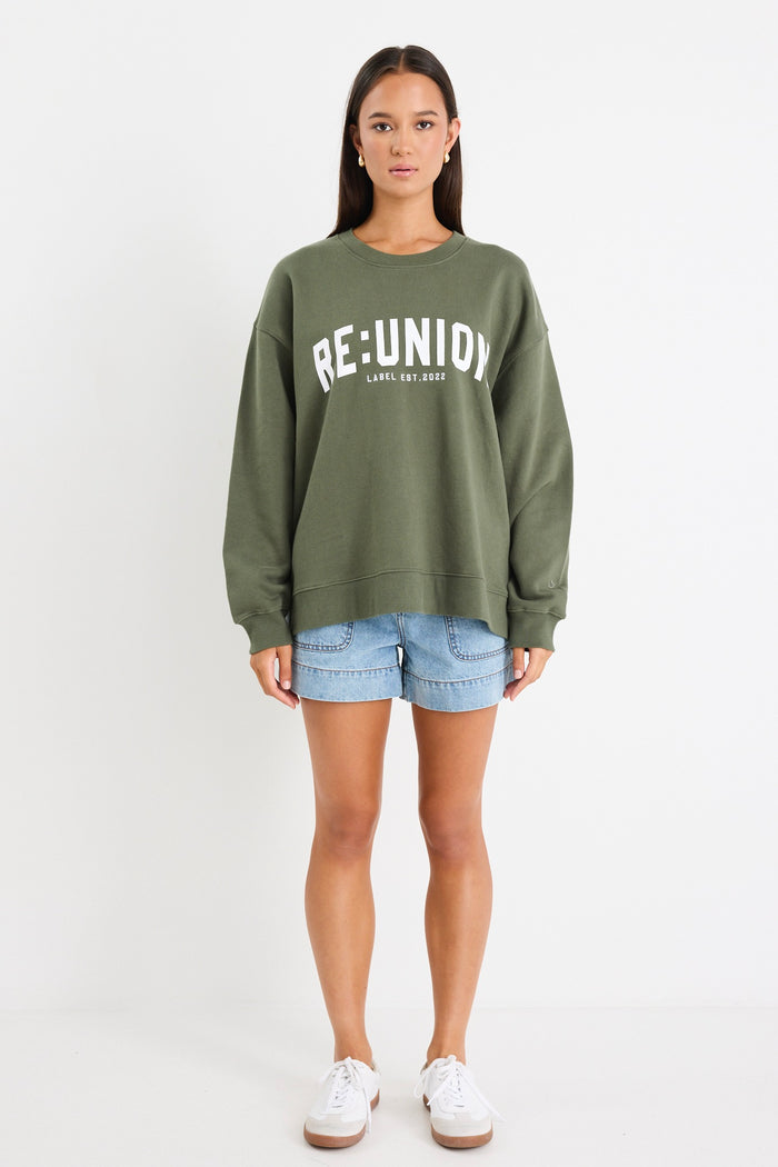 Woman wearing a khaki green oversized sweatshirt with "RE:UNION" printed across the chest, styled with light blue denim shorts.