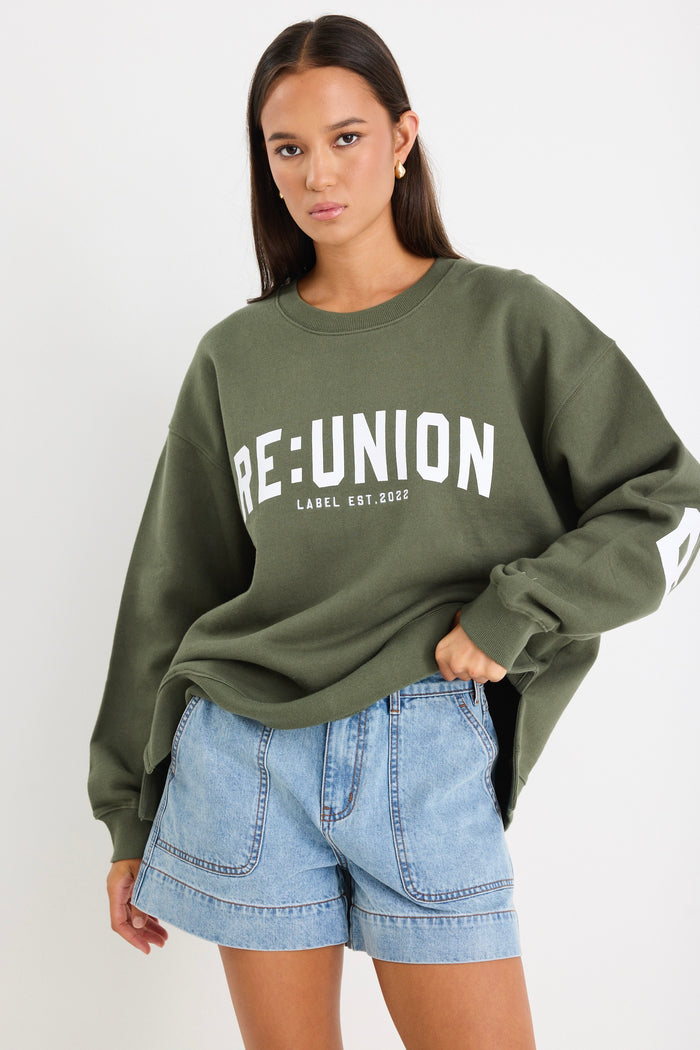 Woman wearing a khaki green oversized sweatshirt with "RE:UNION" printed across the chest, styled with light blue denim shorts.