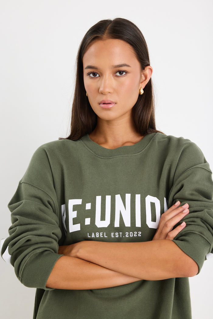 Woman wearing a khaki green oversized sweatshirt with "RE:UNION" printed across the chest, styled with light blue denim shorts.