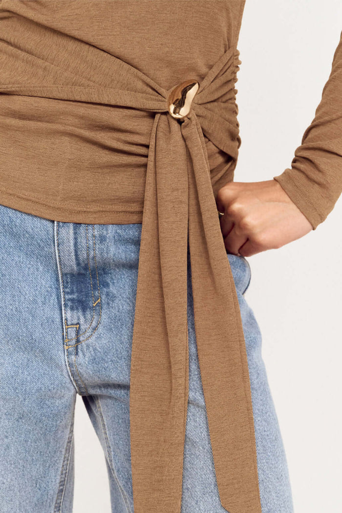 Brown long-sleeve top with a tie detail worn with blue jeans on a plain background