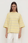 A model wearing a pale yellow oversized short-sleeve t-shirt with thin dark horizontal stripes, paired with white pants, against a plain off-white background.