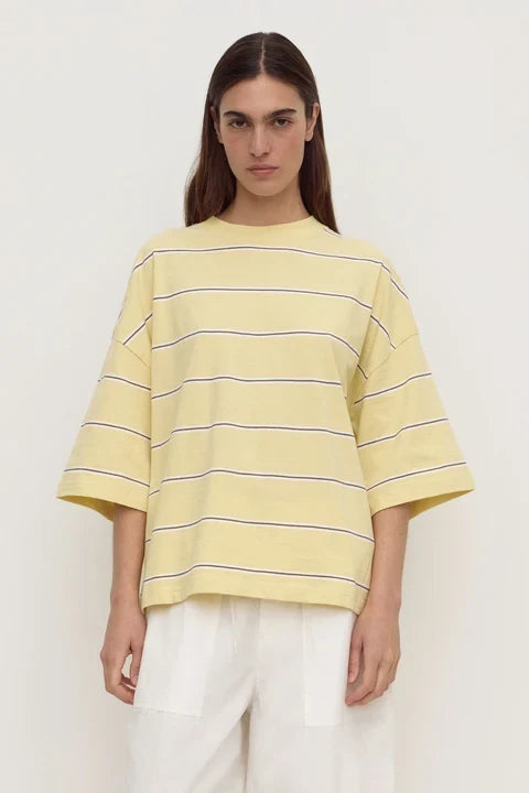 A model wearing a pale yellow oversized short-sleeve t-shirt with thin dark horizontal stripes, paired with white pants, against a plain off-white background.