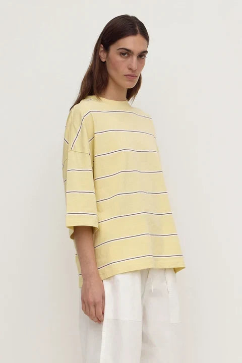 A woman wearing a pale yellow oversized short-sleeve top with thin dark horizontal stripes and white wide-leg pants, photographed against a plain light background.