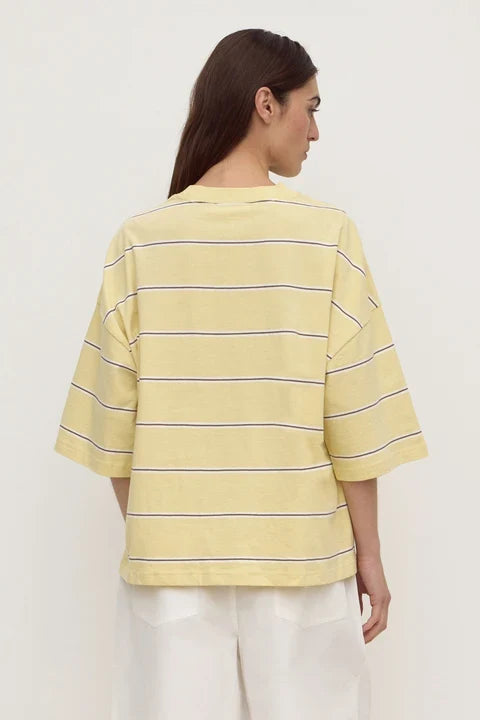 A woman shown from the back wearing a pale yellow short-sleeve striped T-shirt with thin dark horizontal stripes and white pants, photographed against a plain light background.