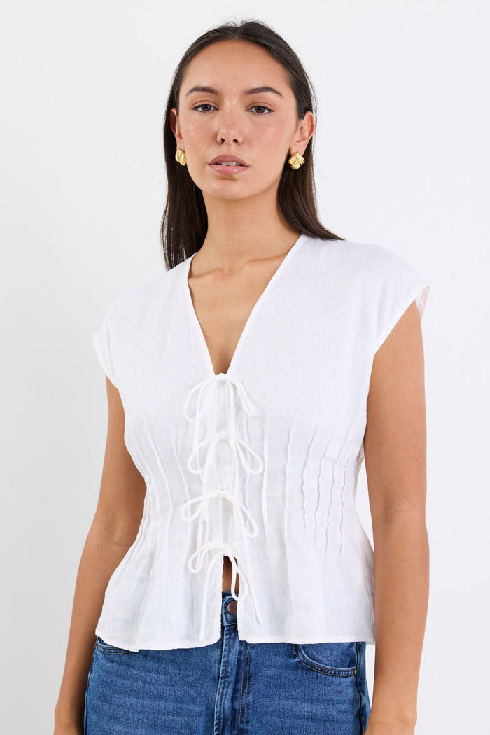Woman wearing an ivory linen cap sleeve vest top with tie-up front detail, paired with blue denim jeans, standing against a white background.