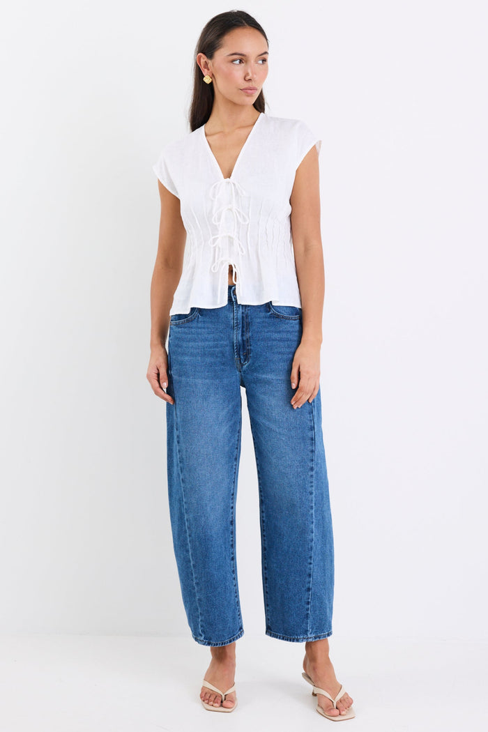 Woman wearing an ivory linen cap sleeve vest top with tie-up front detail, paired with blue denim jeans, standing against a white background.