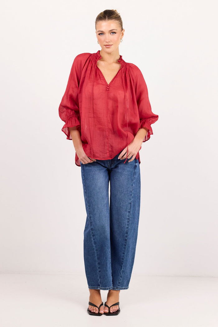 A woman modeling a loose-fitting, long-sleeved red blouse with a gathered neckline, paired with blue jeans and black sandals, standing against a plain white studio background.