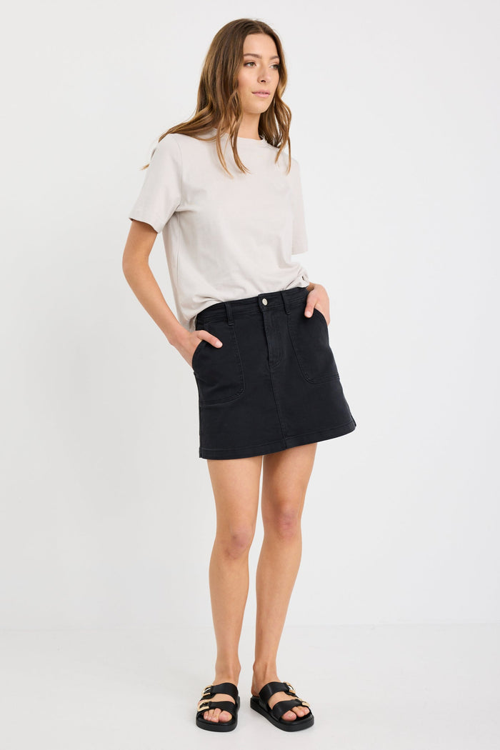 Woman wearing a washed black denim mini skirt with front patch pockets, styled with a tucked-in neutral tee and black sandals.