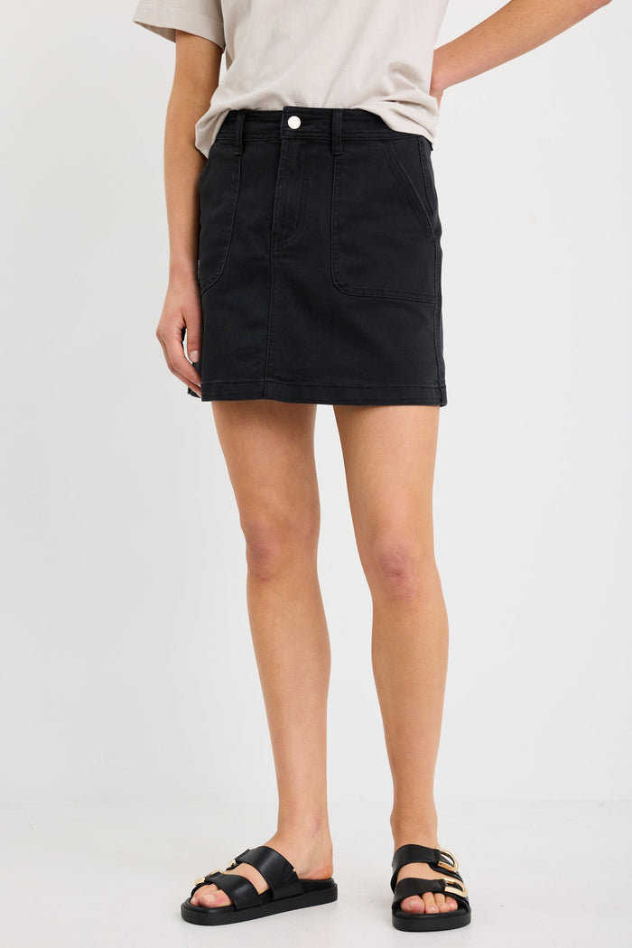 Woman wearing a washed black denim mini skirt with front patch pockets, styled with a tucked-in neutral tee and black sandals.