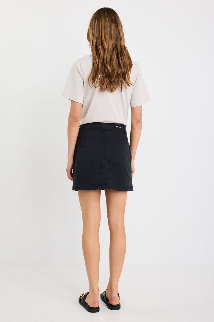 Woman wearing a washed black denim mini skirt with front patch pockets, styled with a tucked-in neutral tee and black sandals.