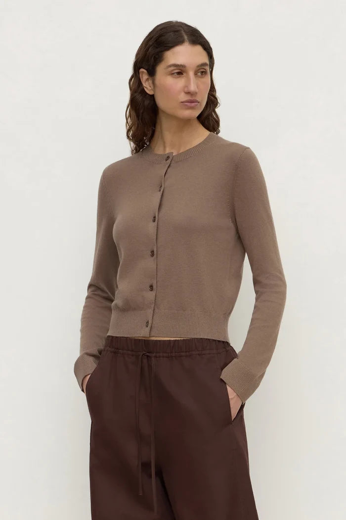 A woman modeling a taupe brown long-sleeve cropped button-front cardigan with dark brown drawstring pants, standing against a plain off-white studio background.