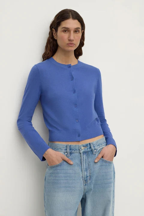 A woman wearing a royal blue long-sleeve cropped button-front cardigan and light-wash blue jeans, standing against a plain light gray wall.