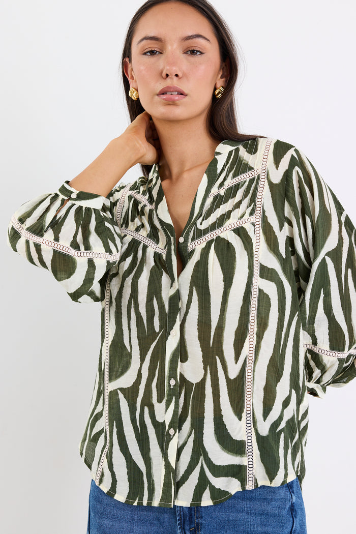 Woman wears a khaki and cream zebra-print shirt with lace inserts and pintuck detail, styled with gold earrings and blue jeans.