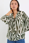 Woman wears a khaki and cream zebra-print shirt with lace inserts and pintuck detail, styled with gold earrings and blue jeans.