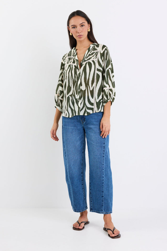 Woman wears a khaki and cream zebra-print shirt with lace inserts and pintuck detail, styled with gold earrings and blue jeans.