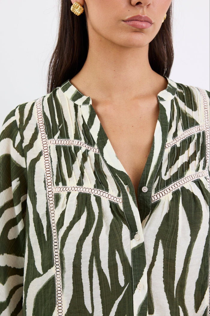 Woman wears a khaki and cream zebra-print shirt with lace inserts and pintuck detail, styled with gold earrings and blue jeans.