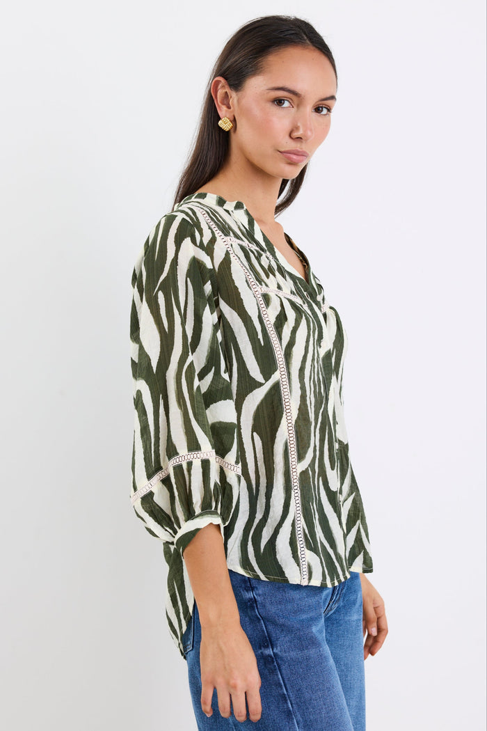 Woman wears a khaki and cream zebra-print shirt with lace inserts and pintuck detail, styled with gold earrings and blue jeans.