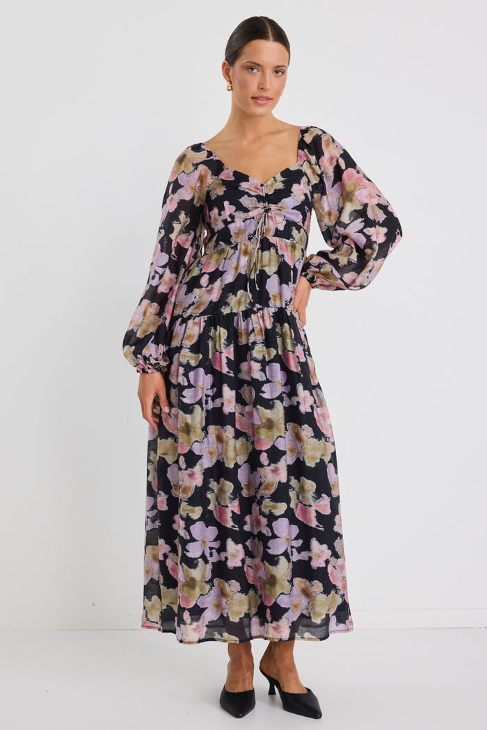 Woman wearing a tiered maxi dress with a bold floral print, ruched front bodice, and long sleeves, styled with black heels.