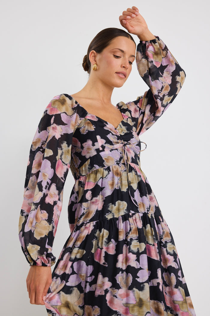 Woman wearing a tiered maxi dress with a bold floral print, ruched front bodice, and long sleeves, styled with black heels.