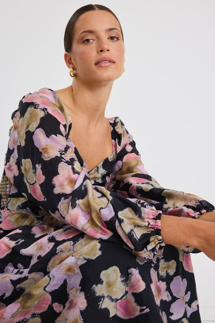 Woman wearing a tiered maxi dress with a bold floral print, ruched front bodice, and long sleeves, styled with black heels.