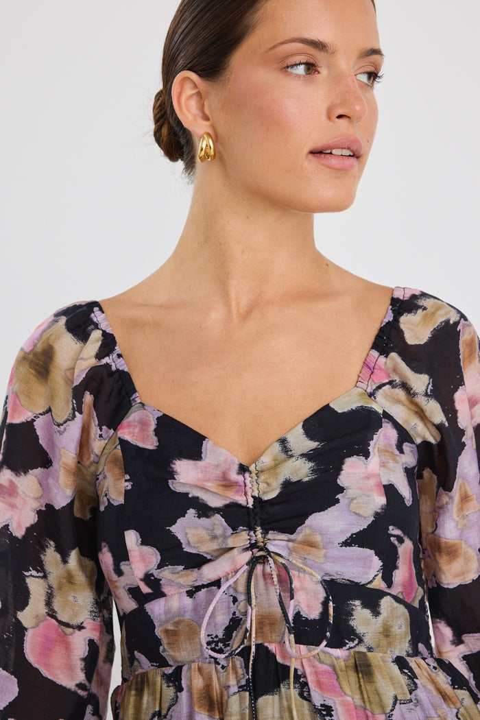 Woman wearing a tiered maxi dress with a bold floral print, ruched front bodice, and long sleeves, styled with black heels.