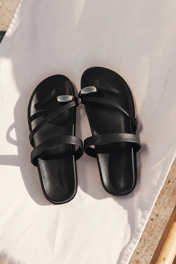 A pair of black leather slide sandals with a small silver metal accent on the front strap, displayed on a white fabric surface.