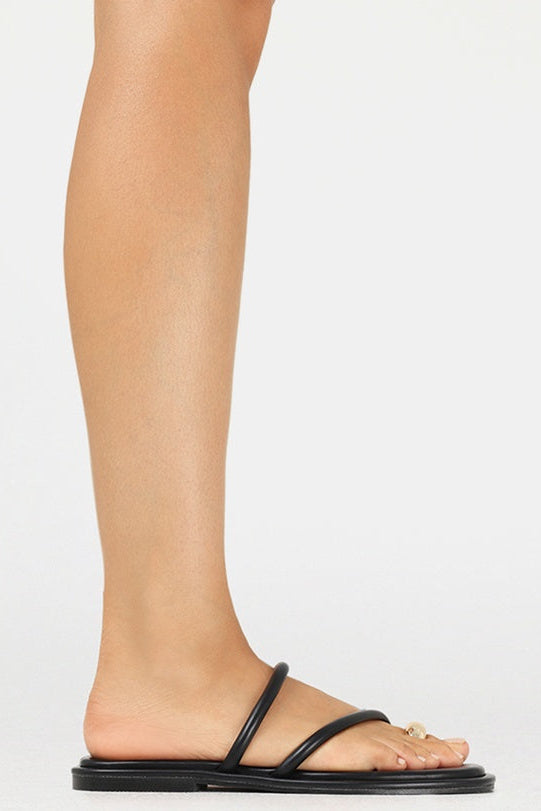 A side view of a person's foot and lower leg wearing a minimalist black flat slide sandal with a single strap and a small gold ring detail on the toe, set against a plain white background.