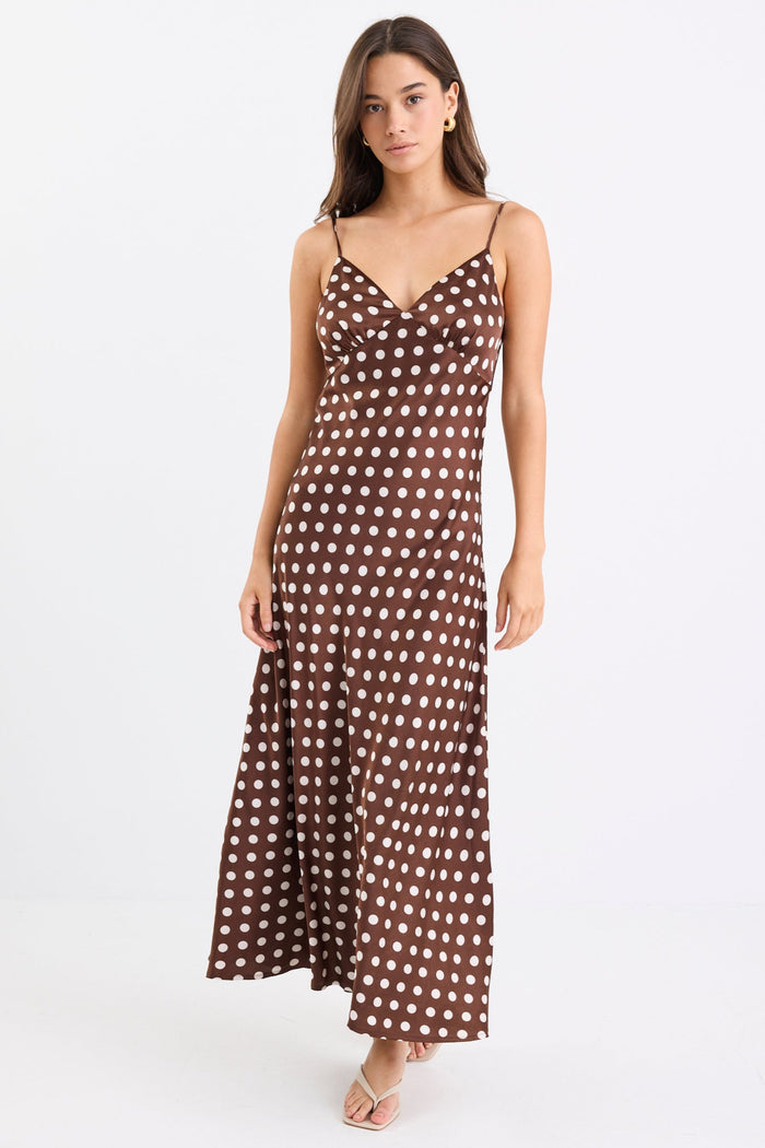 Woman wearing a chocolate brown satin midi dress with white polka dots, adjustable spaghetti straps, and a bias-cut silhouette, standing against a white background.