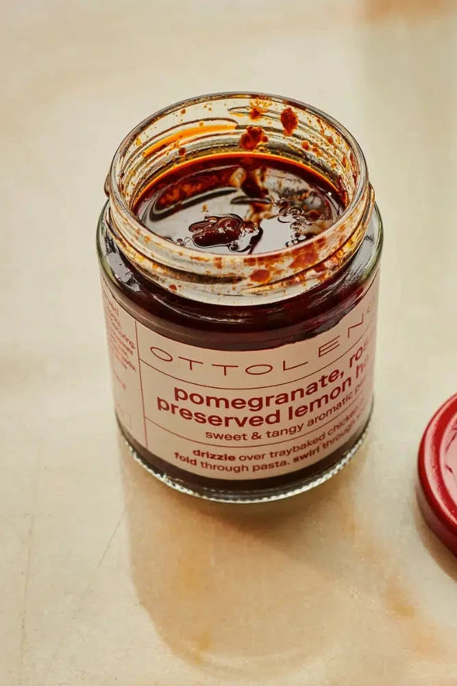 An open jar of Ottolen pomegranate preserved lemon harissa with its red lid placed beside it on a light beige surface. The jar contains a dark reddish-brown paste, and the label reads 'OTTOLEN pomegranate, no preserved lemon harissa sweet & tangy aduki'.