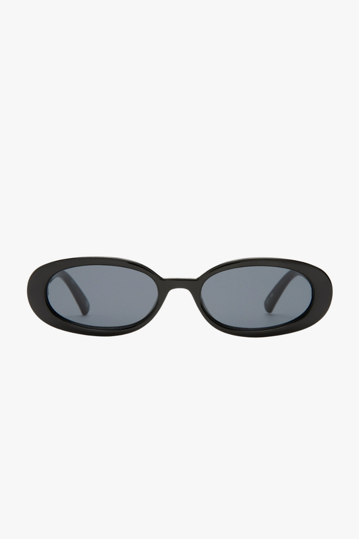 Front-facing view of slim oval sunglasses in a glossy black frame with smoke-tinted lenses. The sleek, narrow silhouette channels a strong 90s influence, offering a bold, minimalist look.