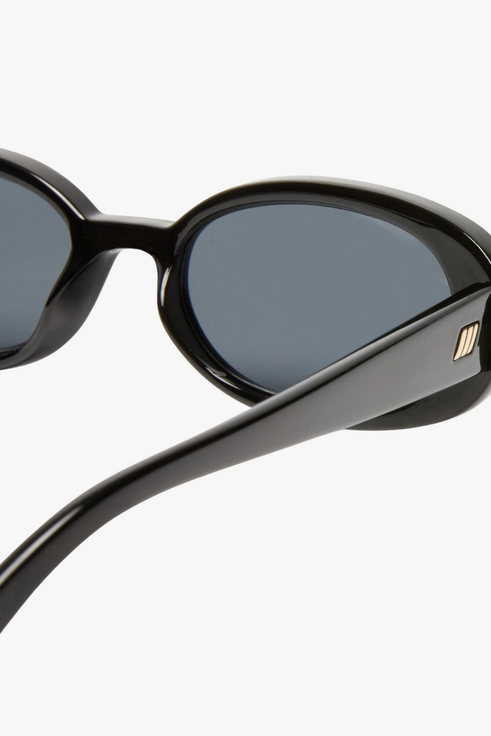 Front-facing view of slim oval sunglasses in a glossy black frame with smoke-tinted lenses. The sleek, narrow silhouette channels a strong 90s influence, offering a bold, minimalist look.