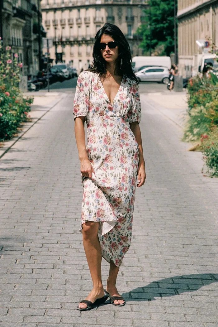 A woman wearing a short-sleeve floral midi dress and black slide sandals walks down a cobblestone city street lined with greenery and buildings.