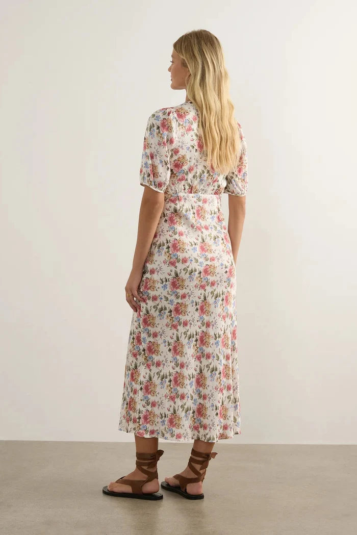 A woman stands with her back to the camera wearing a short-sleeve midi dress with a pink, red, and green floral pattern on a cream background. She is wearing brown strappy sandals in a minimalist studio setting with a light beige wall and concrete floor.