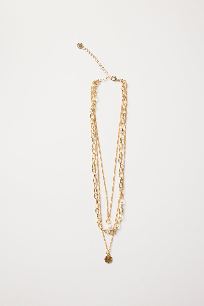 Gold triple-layer necklace with mixed chains, a pearl accent, and a round gold disc charm, styled flat on a white background
