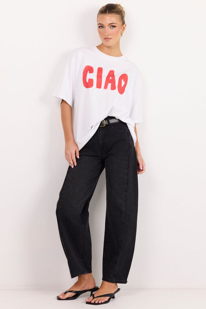 A woman modeling an oversized white t-shirt with 'CIAO' printed in red across the chest, paired with black wide-leg jeans and black sandals, standing against a plain white studio background.