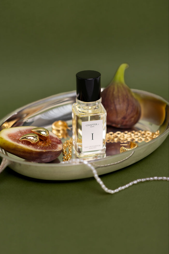 Small bottle of perfume labeled 'I' on a silver tray with figs and jewelry against a green background