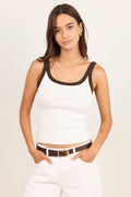 A woman modeling a white ribbed tank top with dark brown trim on the scoop neckline and armholes, paired with white pants and a brown belt, against a plain light beige studio background.
