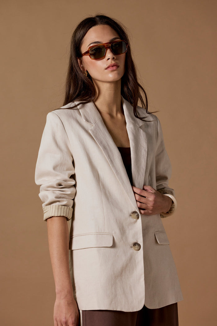 Woman wearing a beige blazer and sunglasses against a brown background
